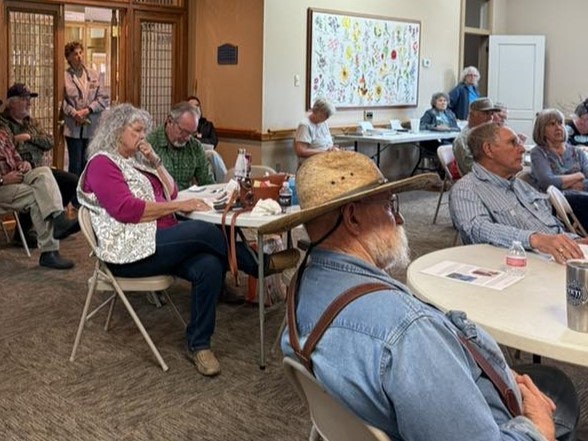 A group of people sitting in a community room, some are listening and others appear engaged in conversation.