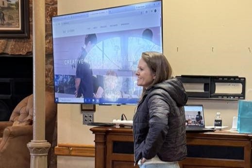 A woman stands in front of a screen displaying a website, with a cozy room in the background featuring wooden furniture.