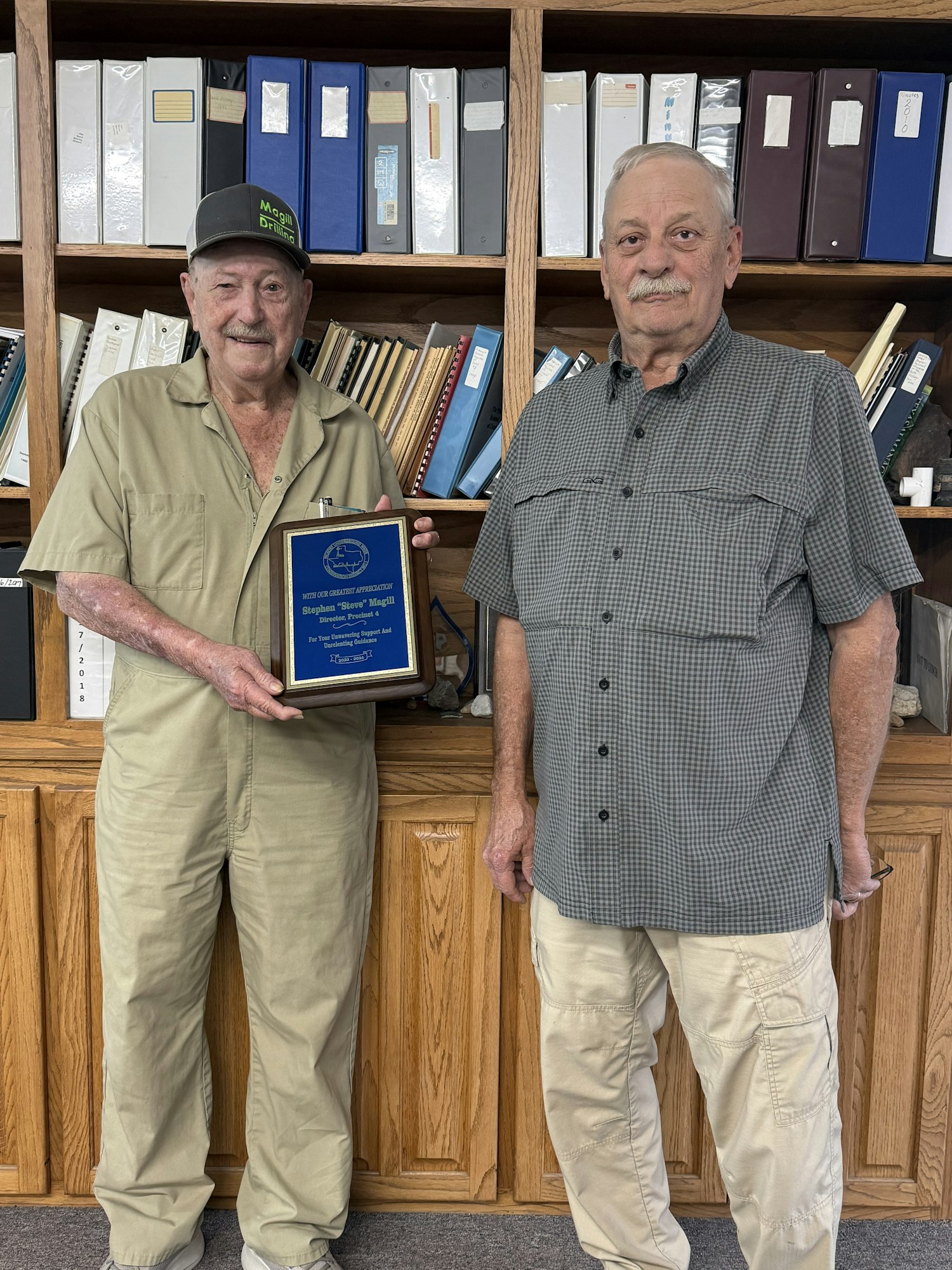 Two men stand together, one holding an award plaque, in front of a bookshelf filled with binders and documents.