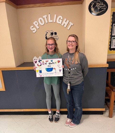 Two people standing indoors, one holding a "Save Water" poster.