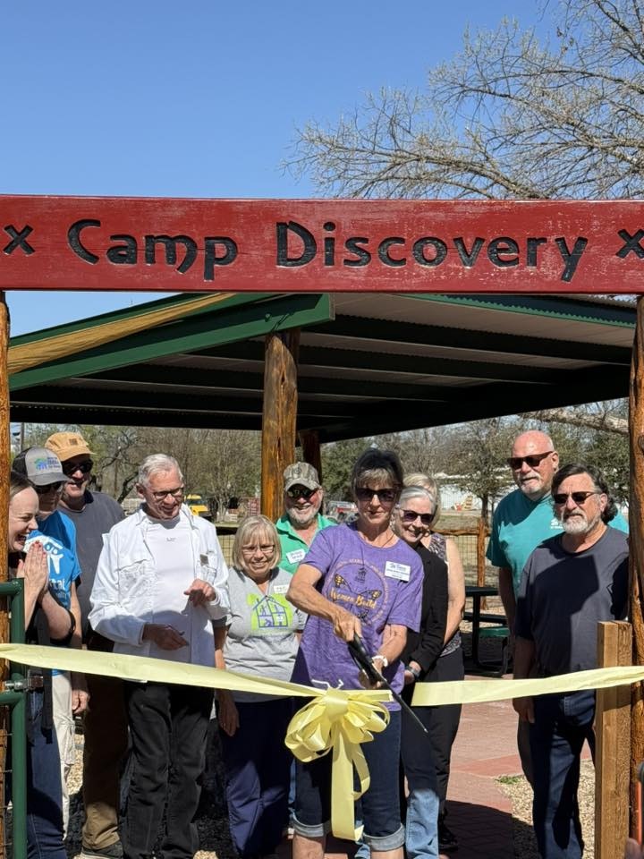 A group of people gathers to celebrate the ribbon-cutting ceremony for Camp Discovery, featuring a yellow ribbon and sign.