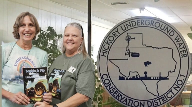 Two people smiling, holding books, stand in front of a window labeled "Hickory Underground Water Conservation District No. 1."