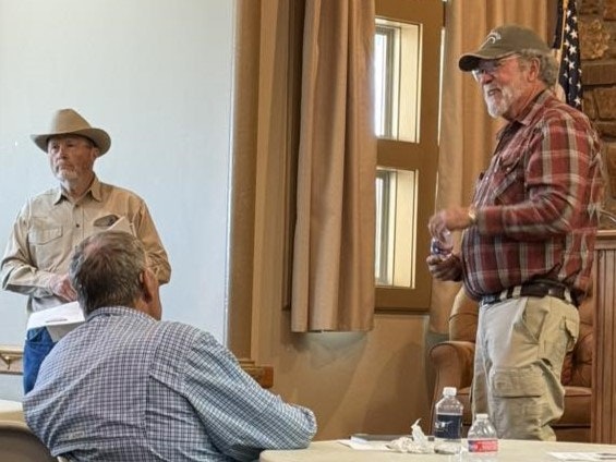 Two men are speaking in front of an audience in a room, with one man engaged in a discussion and another listening.