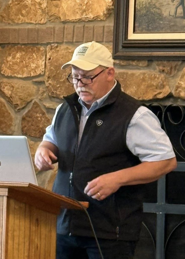 A person stands at a podium with a laptop, wearing a cap and glasses, in front of a stone wall, likely giving a presentation.
