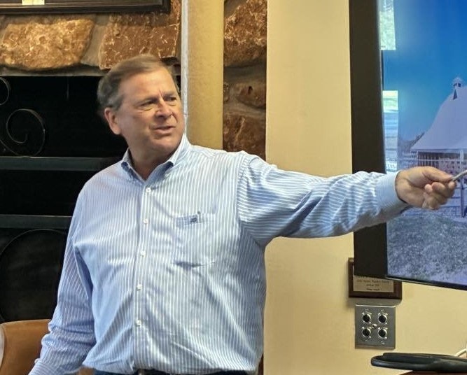 A person gestures while speaking, possibly giving a presentation, with a screen displaying an image in the background.