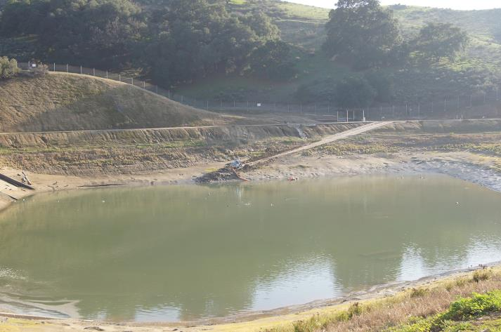 A small pond with surrounding hills and trees under a clear sky.