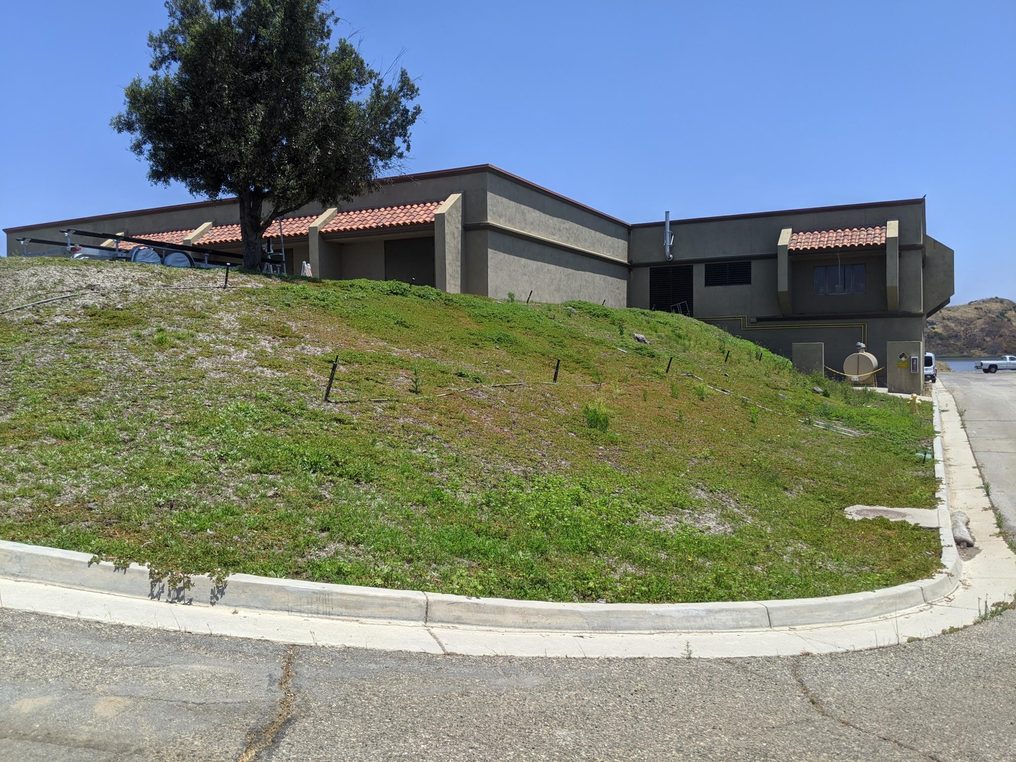 A building with a brown roof, tree, and grass-covered hill, next to a street and body of water in the background.