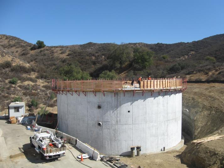 Concrete structure under construction, surrounded by a hillside. Workers and a utility truck visible nearby.