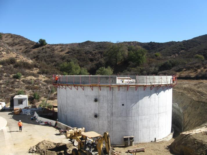 A large cylindrical concrete structure under construction in a hilly area, with construction equipment and workers nearby.