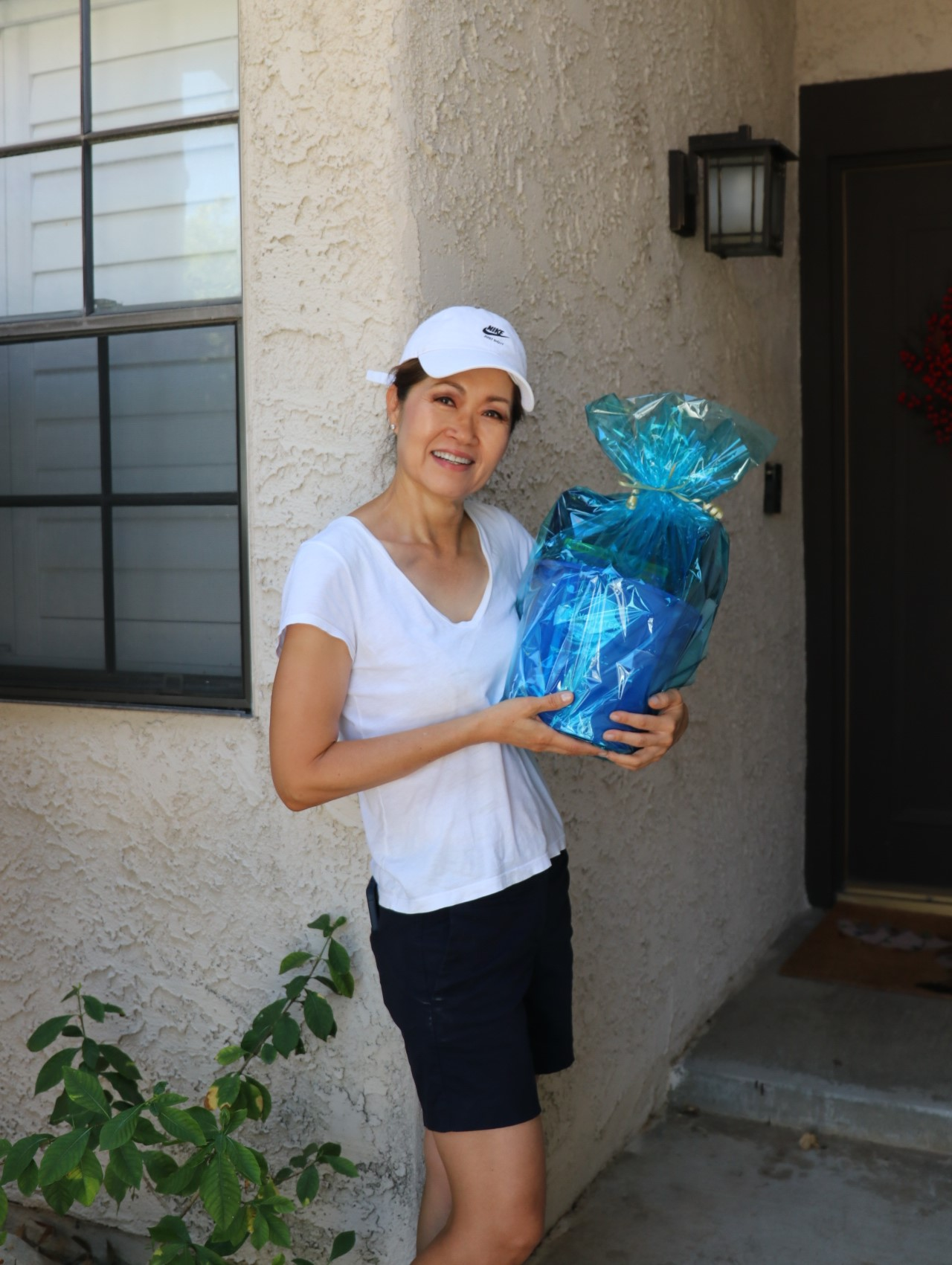 A person in a white hat and shirt holds a blue gift basket on a porch.
