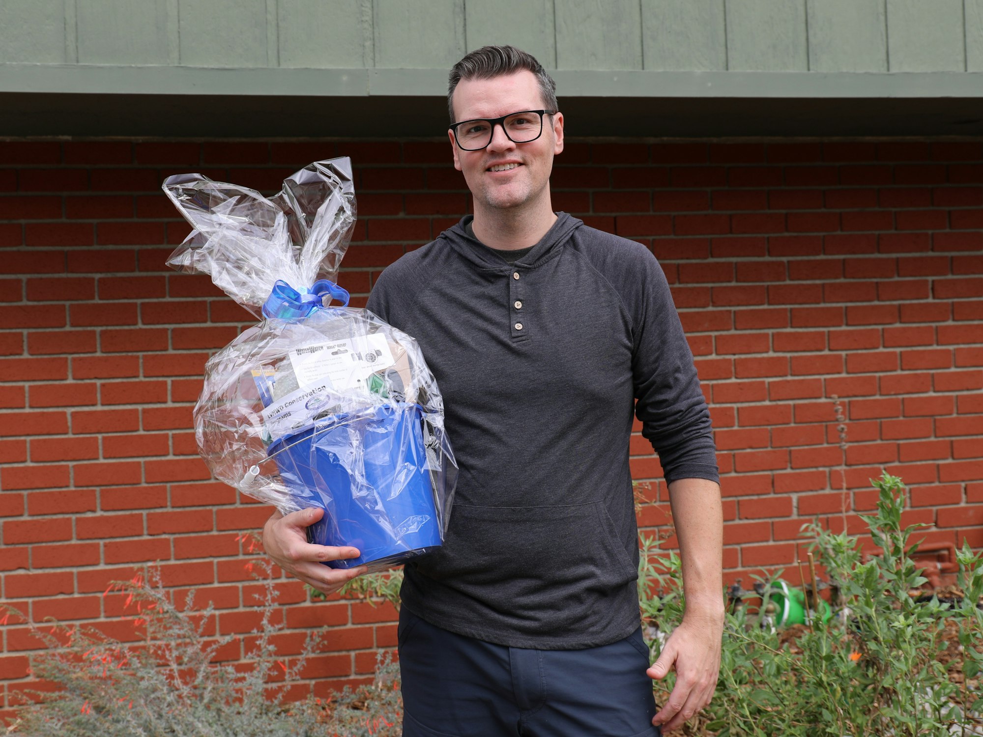 A man holds a wrapped gift basket with a blue container, standing in front of a brick wall with plants around.