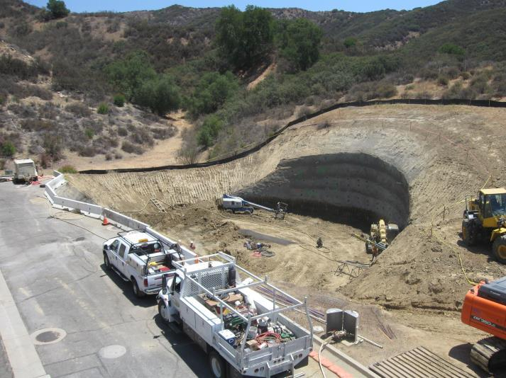 Construction site with trucks, machinery, and a partially excavated hill in a rural area.