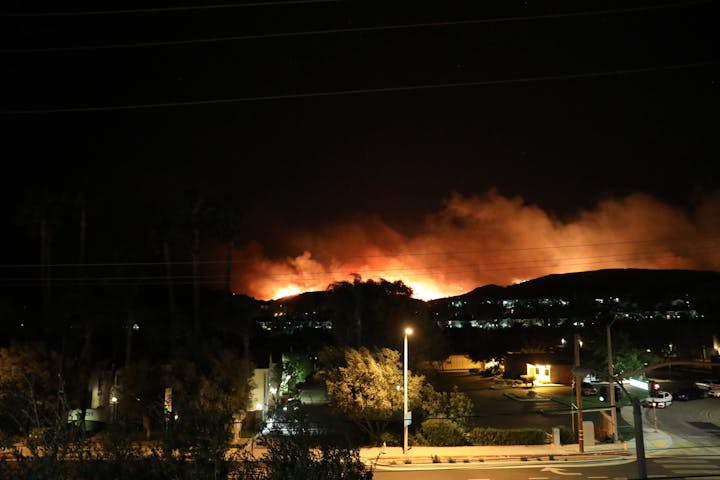 A wildfire is blazing at night over a hill near a town, with smoke rising into the sky.