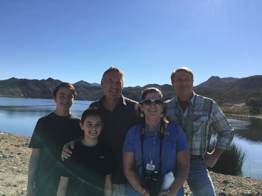A group of people posing by a lake with mountains in the background, under a clear blue sky.