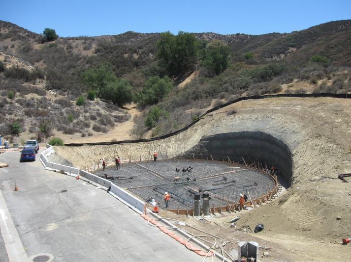 Construction site with workers building a round structure, set in a hilly landscape with a parked car and safety barriers.