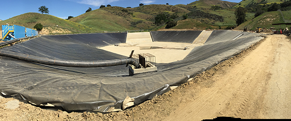 A large, empty, lined pond structure with surrounding dirt pathways and hills in the background.