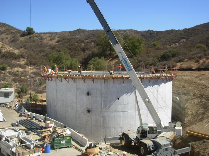 Construction site with a large cylindrical concrete structure, crane, and workers, set in a hilly landscape.