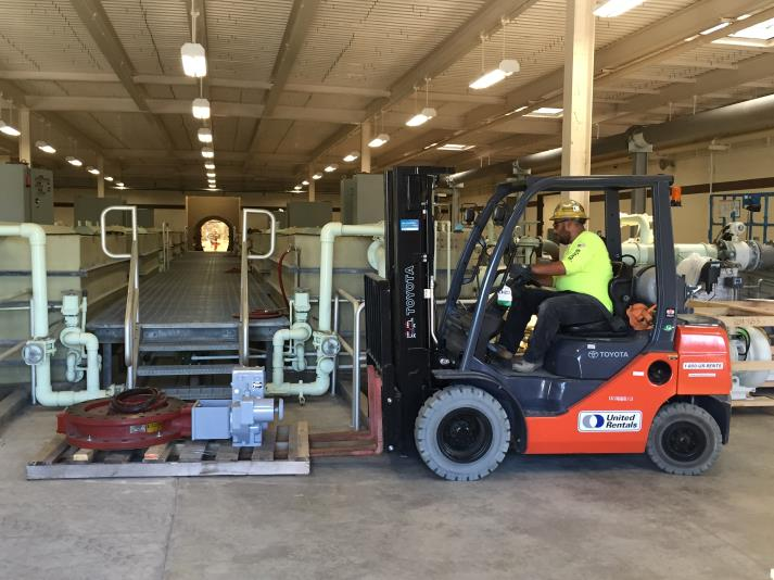 A worker operates a forklift to move equipment on a pallet in an industrial setting with pipes and machinery.