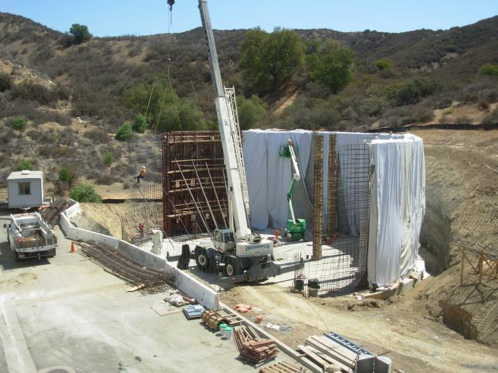 Construction site with a crane lifting materials, reinforced walls being built, and workers on lift equipment, set in a hilly area.