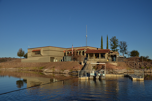A building with a red-tiled roof by a lake or river, surrounded by trees, under a clear blue sky.