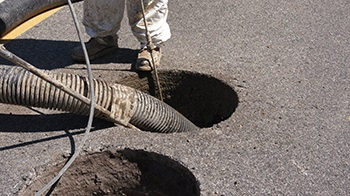 A worker using a hose to clean or inspect a hole in the ground on a paved surface.