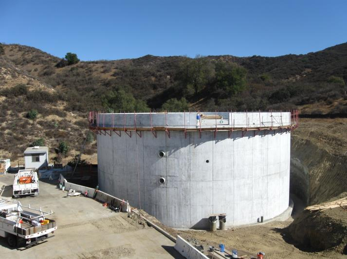 A large concrete structure, possibly a tank, with vehicles and construction materials nearby, set in a hilly area.