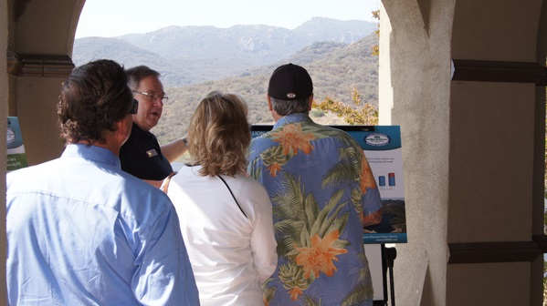 A group of people are looking at informational boards in a mountainous, outdoor setting.