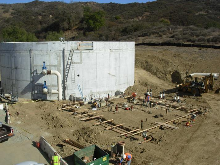 A construction site with a large cylindrical concrete structure, workers, and a bulldozer in a hilly area.