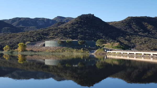 A calm lake reflects a hilly landscape, trees, and a building with a clear blue sky.
