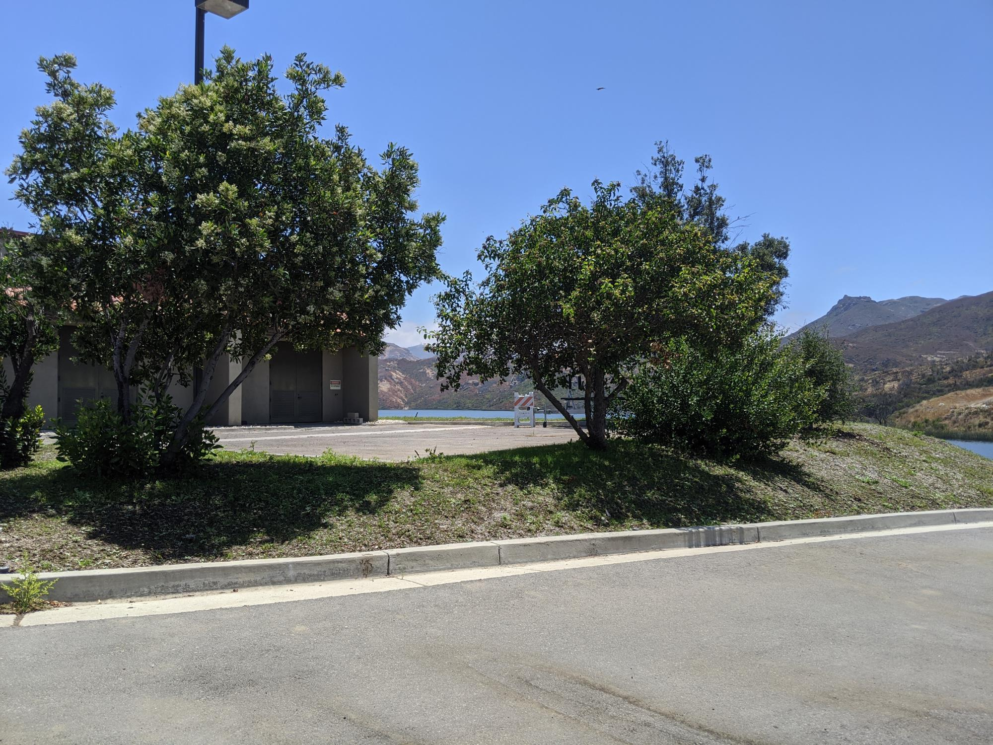 Landscape with trees, a building, mountains in the background, blue sky, and a paved area in the foreground.