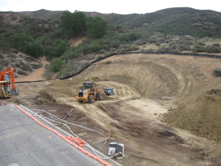 Construction site with earth-moving equipment in a hilly landscape, featuring a backhoe and a bulldozer on dirt terrain.