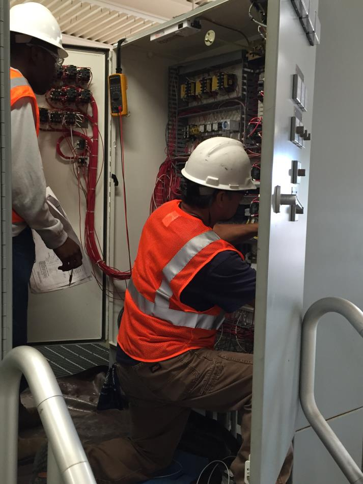 Two workers in safety gear inspecting electrical cabinet wiring.