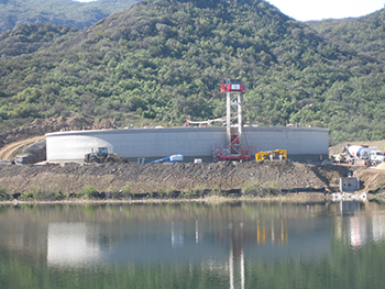 A circular structure under construction near a body of water, with hills in the background.