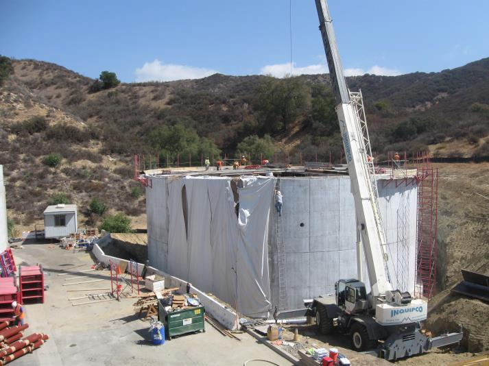 A construction site with a large cylindrical structure, surrounded by scaffolding and a crane, in a hilly landscape.