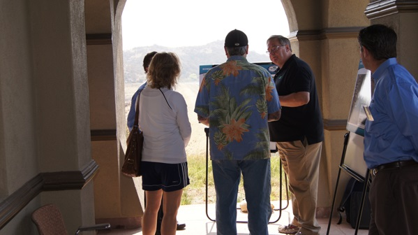 A group of people talking in an archway, one person gesturing, outdoor setting with hills in the background.
