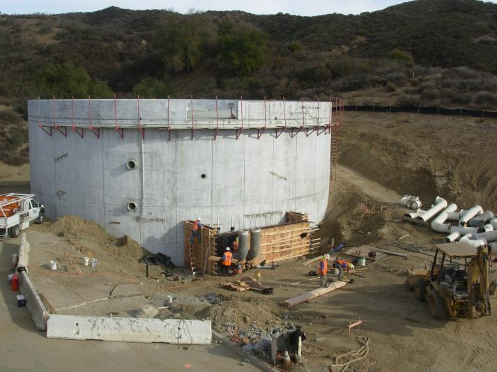 A construction site with a large circular concrete structure, workers, machinery, and pipe materials.