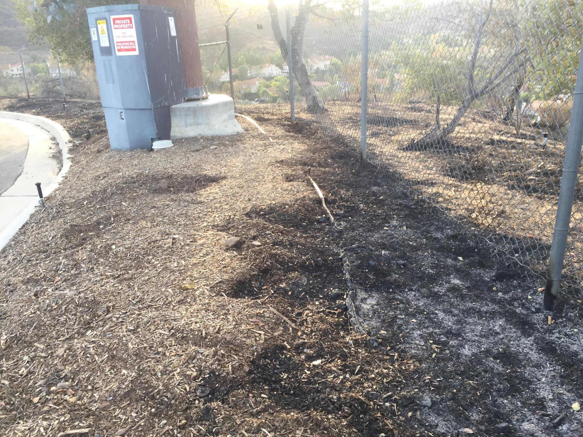 A dirt path with burnt patches, a fence, a utility box with warning signs, and a tree on a hill.
