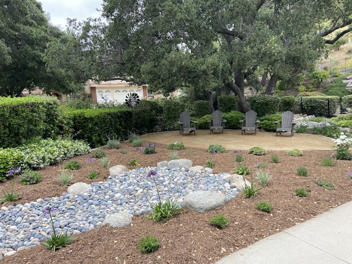 A landscaped garden features a pebble stream, seating area with chairs, and various plants, surrounded by lush greenery.