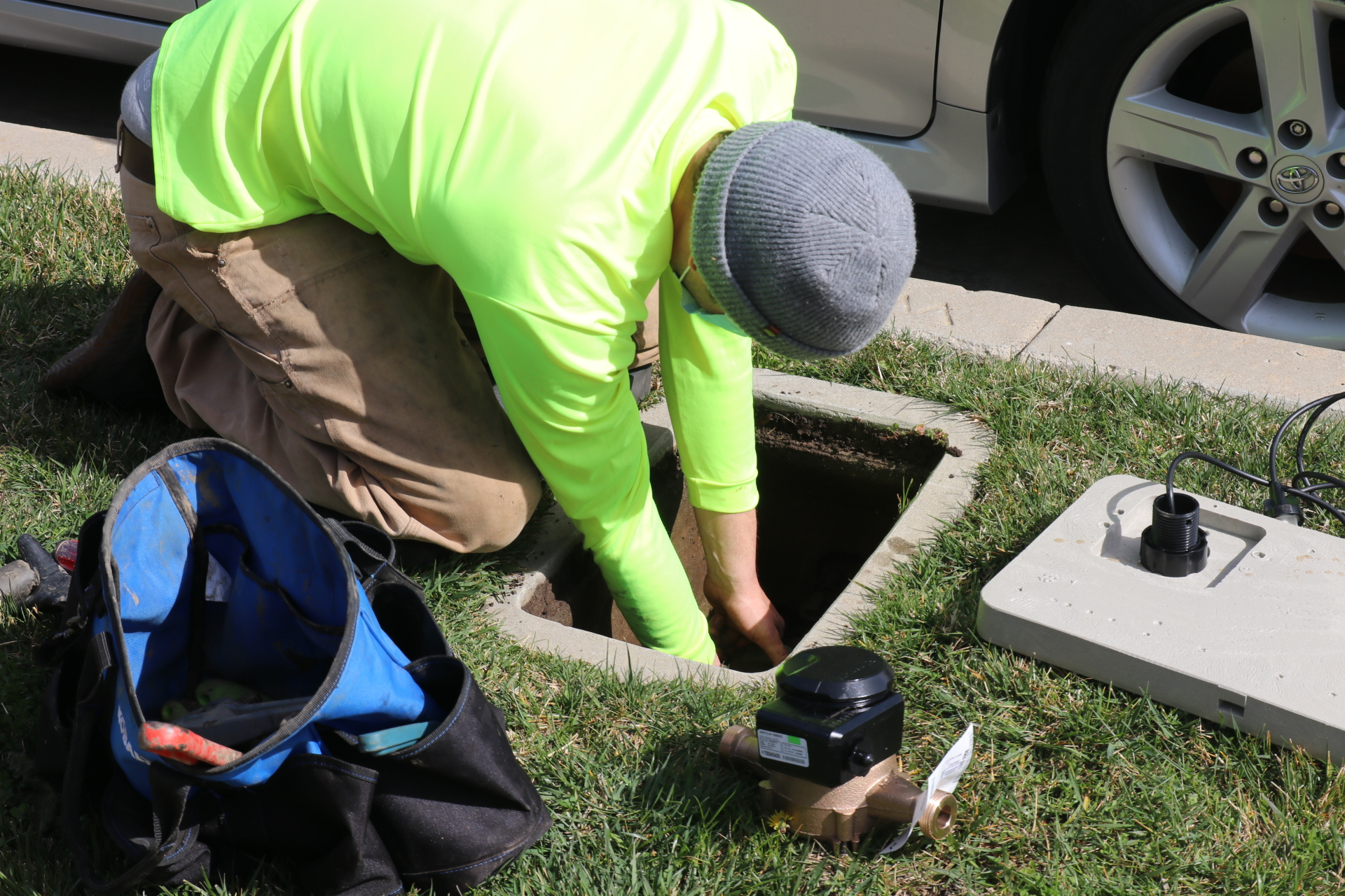 A person in a bright shirt and beanie works in a utility box by the roadside. A tool bag and equipment are nearby.