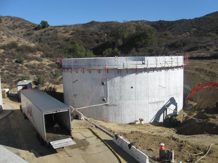 A large cylindrical concrete structure under construction, with a truck and excavator nearby, set in a hilly area.