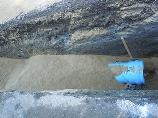 A trench with sand at the bottom, containing a partially buried blue object, possibly a pipe or bucket.