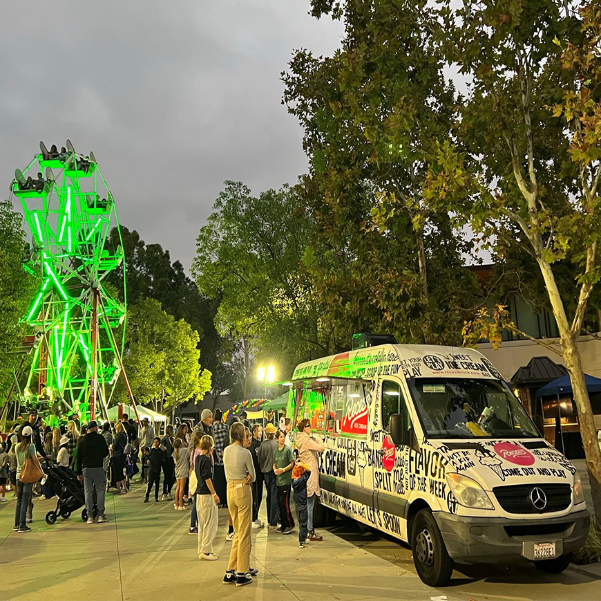 Crowd by food truck at night with a lit Ferris wheel and trees nearby.
