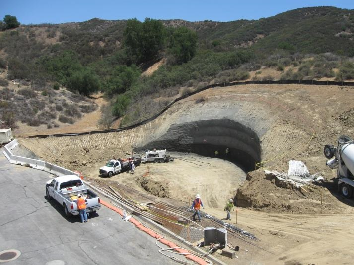 Construction site with workers, vehicles, and an excavated tunnel entrance in a hilly landscape.