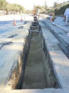 A long trench under construction beside a road, with an excavator, cones, and a ladder.