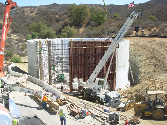 Construction site with cranes, workers, and concrete forms onsite, surrounded by hills and trees.