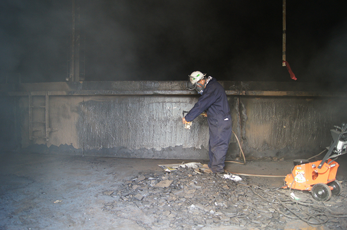 A person in protective gear uses equipment inside an industrial setting, with debris on the floor and machinery nearby.