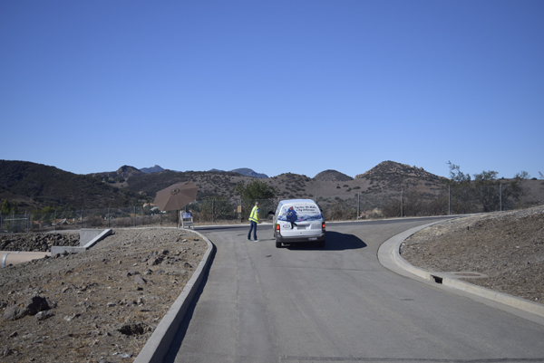 A person in a safety vest stands near a white van on a curved road in a mountainous, dry landscape under a clear blue sky.