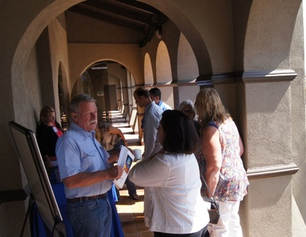 A group of people stand talking under an outdoor arcade with archways, one holding a piece of paper.