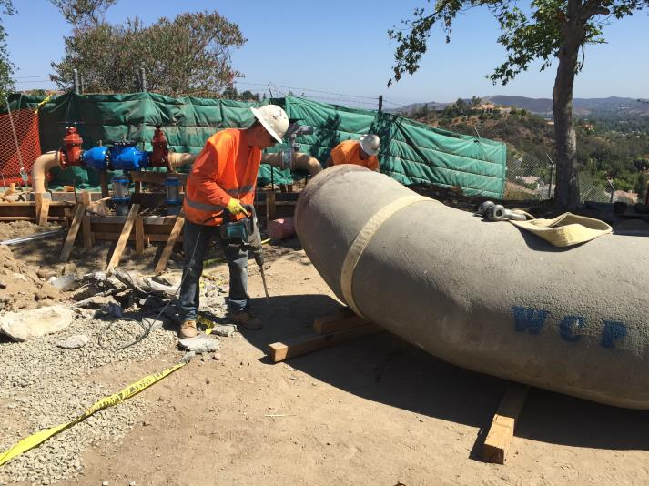 Construction workers in safety gear handling a large concrete pipe at a worksite with equipment nearby.
