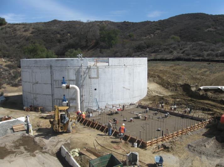 Construction site with a large cylindrical tank, workers, a backhoe, and hills in the background.